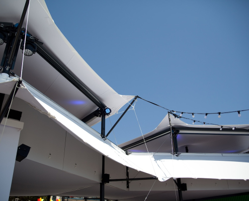 Underside view of stretch tent canopy roofing showing stage lighting rigs and string lights against a blue sky