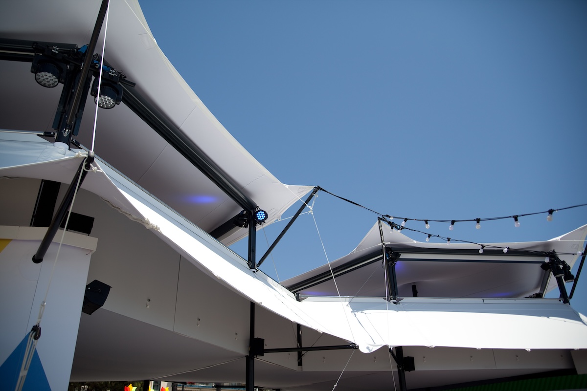 structure-canopy-underside-stage-lighting-rigging-daytime Underside view of stretch tent canopy roofing showing stage lighting rigs and string lights against a blue sky