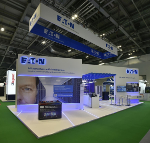 Eaton exhibition stand - full stand overview Full overview of the Eaton exhibition stand featuring the 'Infrastructure with Intelligence' branded wall, hanging Eaton signage and interactive game station