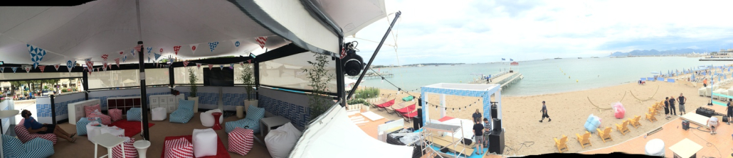 HEXIS structures – beachfront lounge setup Panoramic view of a HEXIS beach lounge structure with striped seating and bunting, overlooking a seafront and pier