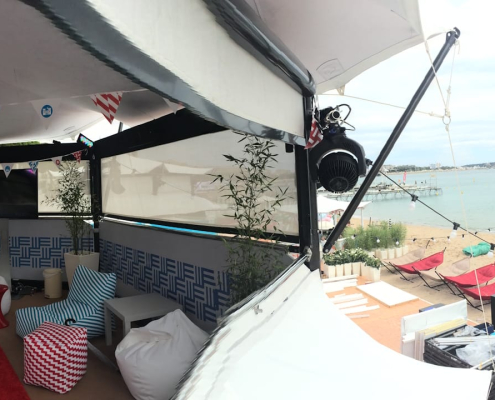 Panoramic view of a HEXIS beach lounge structure with striped seating and bunting, overlooking a seafront and pier