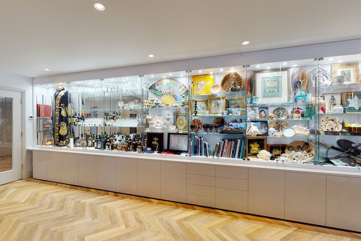 Wooden glass display cases – private collection display wall Long glass display cabinet with grey storage base showing an eclectic collection of glassware, ceramics, shells, books and a decorative robe