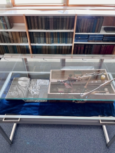 Sabhal Mòr Ostaig glass-topped display case Glass-topped table display case at Sabhal Mòr Ostaig with navy blue velvet lining, showing an open book with a black and white portrait and a wooden box of historic artefacts, in front of library bookshelves