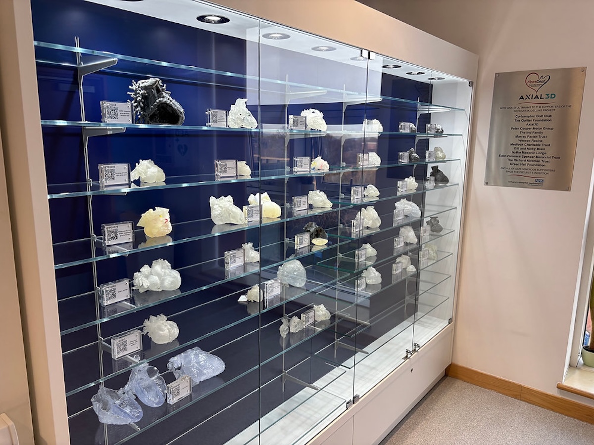 Bespoke glass display cabinet for 3D-printed heartsÛ Bespoke wall-mounted glass display cabinet with navy blue backdrop, illuminated glass shelves and white 3D-printed heart models, beside an Axial3D donor plaque at Southampton hospital