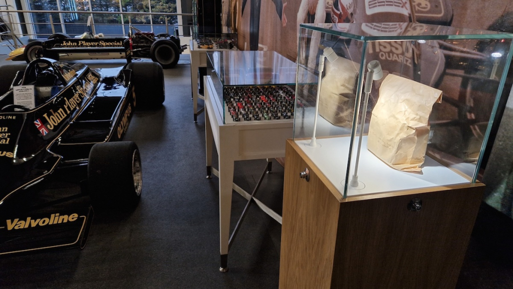 Classic Team Lotus bespoke glass display cabinet Classic Team Lotus bespoke glass display cabinet on an oak base with white LED spotlights, housing Hazel Chapman’s sealed envelope in a paper bag, beside black and gold John Player Special Lotus F1 cars