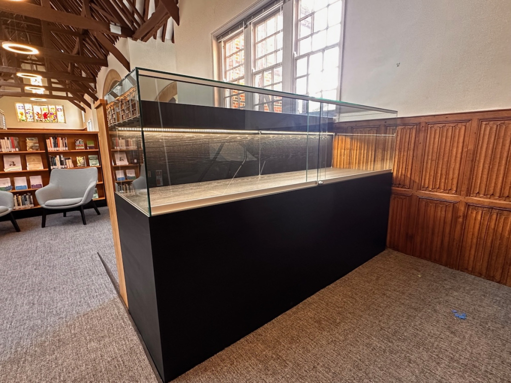 Radley College bespoke glass display cabinet Bespoke glass display cabinet at Radley College with a black base, dark wood-grain interior and warm LED strip lighting, in a library with exposed timber roof beams and carved oak panelling