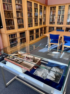 Sabhal Mòr Ostaig glass display table in the library Glass-topped display table with navy blue velvet lining at Sabhal Mòr Ostaig, showing a wooden box of instrument-like artefacts and a black and white photograph, beside light oak glazed bookcases and blue chairs in the library
