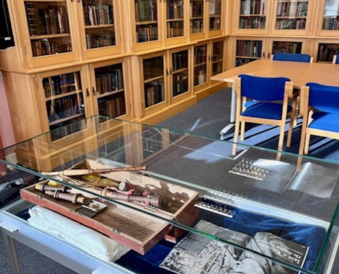 Glass-topped display table with navy blue velvet lining at Sabhal Mòr Ostaig, showing a wooden box of instrument-like artefacts and a black and white photograph, beside light oak glazed bookcases and blue chairs in the library