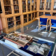 Glass-topped display table with navy blue velvet lining at Sabhal Mòr Ostaig, showing a wooden box of instrument-like artefacts and a black and white photograph, beside light oak glazed bookcases and blue chairs in the library