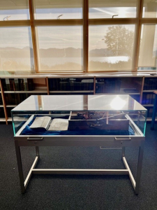 Sabhal Mòr Ostaig glass-topped display table Free-standing glass-topped display table with a silver metal frame and navy blue velvet lining at Sabhal Mòr Ostaig, displaying an open book and artefacts in front of library windows overlooking a loch and hills
