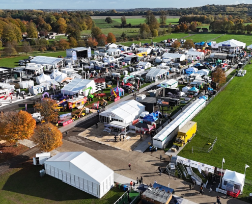 Aerial view of an outdoor trade show on a green showground in autumn, with white marquees, event structures and exhibitor stands set among orange and gold trees