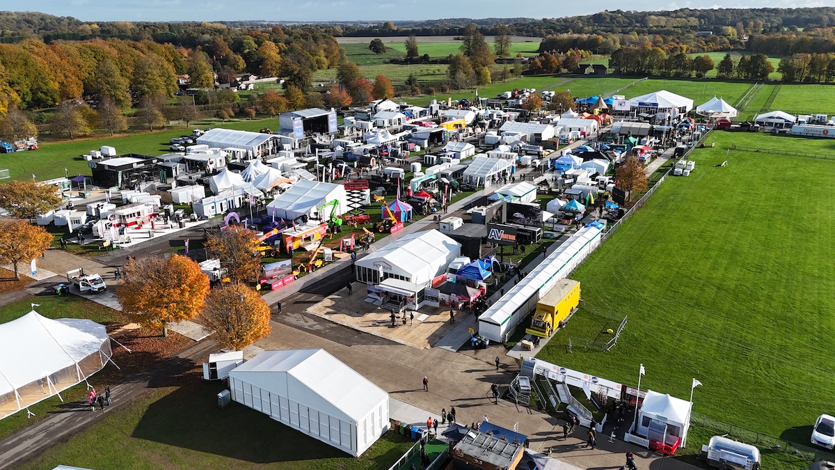 Aerial view of an outdoor trade show on a green showground in autumn, with white marquees, event structures and exhibitor stands set among orange and gold trees