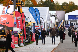 Visitors walking along a wooden boardwalk at an outdoor trade show in autumn, past red and blue teardrop flags, yellow JCB telehandlers and white marquees