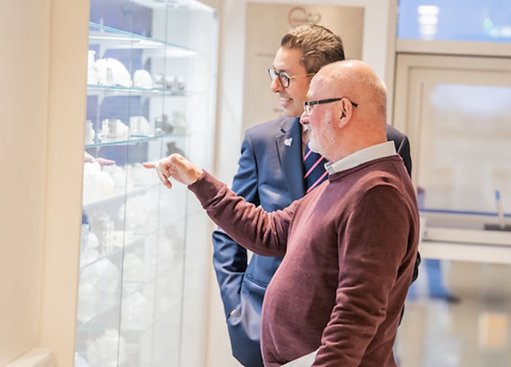 Viewing the 3D-printed hearts display cabinet Two men in glasses, one in a navy blazer and one in a burgundy jumper, looking at 3D-printed heart models on illuminated glass shelves in a bespoke display cabinet